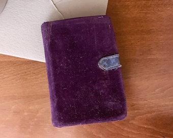 May include: A rectangular, deep purple velvet box with a small, faded blue clasp. The box is slightly worn, suggesting age. The background includes a light-colored envelope and a wooden surface.