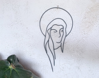 May include: A minimalist metal wall art piece depicting a woman's face with a halo. The artwork is crafted from a single, continuous line of dark metal and is mounted on a textured white wall. The piece has a simple, elegant design.