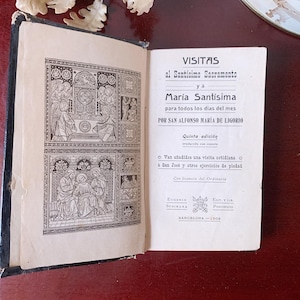 May include: An open antique book with black and white illustrations and text. The book's title page reads "VISITAS al Santisimo Sacramento y a Maria Santisima". The book is on a dark red surface.