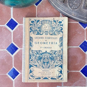 Peut inclure: Un livre ancien intitulé "Lecciones Elementales de Geometría" en espagnol, avec des motifs bleus et blancs. Le livre est légèrement usé et repose sur une surface carrelée avec des motifs géométriques roses et bleus.
