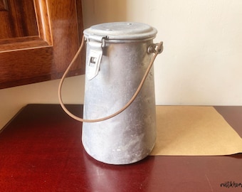 May include: A vintage metal milk can with a lid and a wire handle. The can is silver with a brown handle and a latch closure. The can is sitting on a dark red surface.