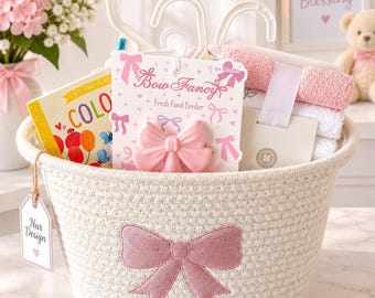 Può includere: Un cesto in corda intrecciata bianca con un fiocco rosa ricamato sul davanti e un'etichetta Nur Design. All'interno si trovano un ciuccio per alimenti Bow Fancy, un libro colorato con alette e salviette per neonati piegate.