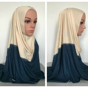 May include: A two-piece hijab in beige and navy blue. The beige top section has delicate embellishments. The lower part is a solid navy blue. The hijab is draped over a mannequin head, displaying its design and fit.
