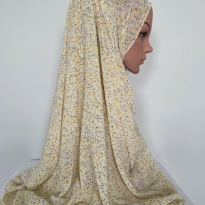 Op de afbeelding: Een lichtgele hijab met kleine gele en grijze bloemmotieven. De stof is over een mannequinhoofd gedrapeerd, waarbij het ontwerp en de textuur van de hoofdbedekking worden benadrukt.
