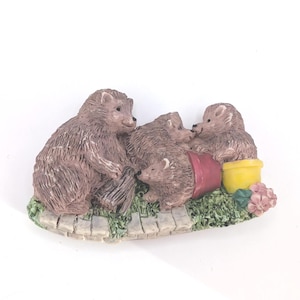 May include: A decorative figurine of a hedgehog family. The brown hedgehogs are gathered around a red flower pot and a yellow pot. The base is a stone path with green grass and pink flowers. A charming garden scene.