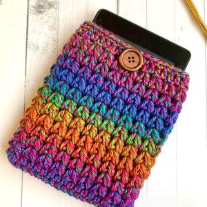 May include: A multicolored crochet sleeve featuring a vibrant gradient of blue, purple, pink, orange, and yellow yarn. The rectangular pouch has a textured stitch pattern and is secured with a single round brown wooden button at the top center.