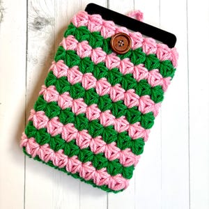 May include: A handmade crochet phone case in a vibrant pink and green pattern. The case features a button closure and is designed to hold a phone. The case is made of soft yarn and is a unique accessory.