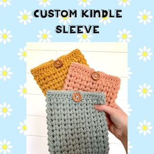 May include: Three crocheted Kindle sleeves in different colors: mustard yellow, peach, and gray. Each sleeve has a wooden button closure. The text "CUSTOM KINDLE SLEEVE" is displayed at the top of the image.