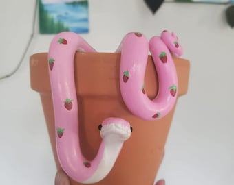 Puede incluir: Una serpiente de cerámica rosa con diseños de fresas está envuelta alrededor de una maceta de terracota.