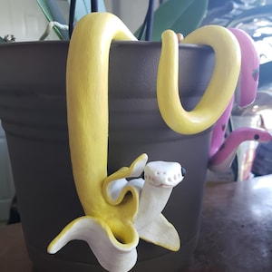 Op de afbeelding: Een gele bananen-sculptuur met een witte schil, geplaatst in een bruine bloempot. De banaan is om de pot gewikkeld en heeft een glimlachend gezicht.