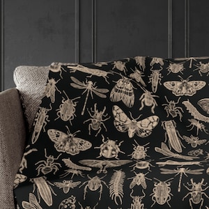 May include: A black throw blanket featuring a detailed beige illustration of various insects, including butterflies, beetles, and dragonflies. The blanket is draped over a gray sofa, showcasing the intricate insect pattern.