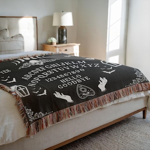 May include: Black woven throw blanket with a white Ouija board design. The blanket features the alphabet, numbers, and phrases like "Hello" and "Goodbye." Decorative fringe in shades of brown and red lines the edges. The blanket is displayed on a bed.