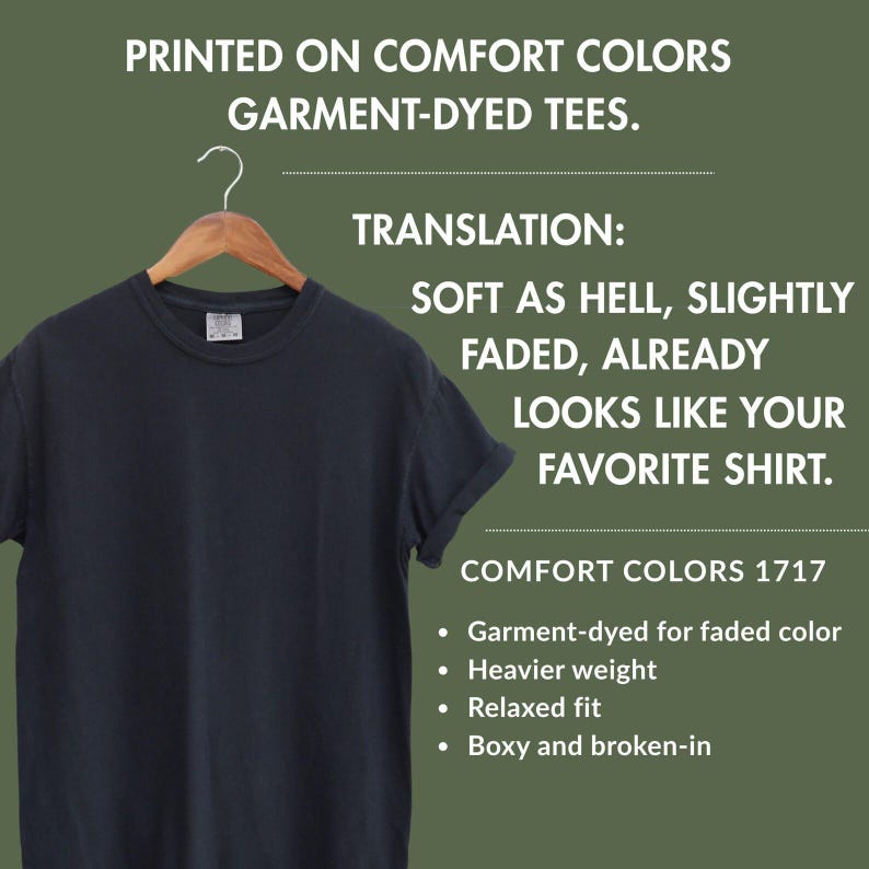 May include: A black short-sleeved t-shirt on a wooden hanger. Text reads: PRINTED ON COMFORT COLORS GARMENT-DYED TEES. TRANSLATION: SOFT AS HELL, SLIGHTLY FADED, ALREADY LOOKS LIKE YOUR FAVORITE SHIRT. COMFORT COLORS 1717. Garment-dyed, heavier weight, relaxed fit.