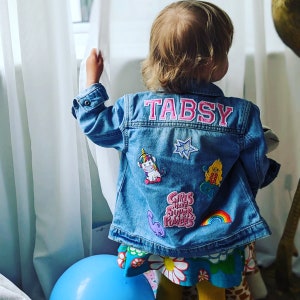 May include: A light blue denim jacket with the name "TABSY" in pink letters on the back. The jacket is decorated with colourful patches, including a unicorn, star, mermaid, dinosaur, and rainbow. The text "GIRLS HAVE SUPER POWERS" is also visible.