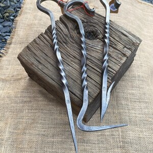 May include: Three hand-forged metal tools with twisted handles and curved or pointed ends. The tools are silver-colored and rest on a weathered wooden block and burlap fabric. Each tool has a small tag attached with red string.