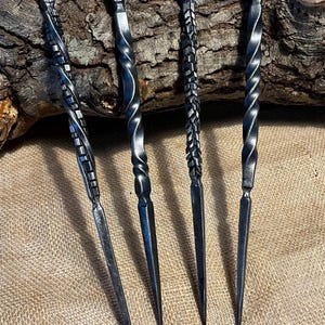 Puede incluir: Cinco pinchos de metal negro con mangos retorcidos. Los pinchos están dispuestos sobre un trozo de madera.
