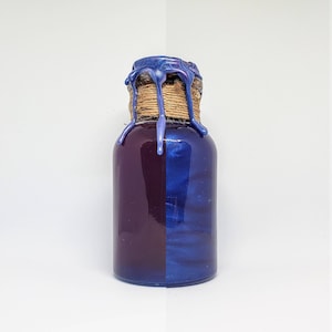 May include: A small, glass bottle with a two-tone color scheme: deep purple on the left and shimmering blue on the right. The bottle's neck is wrapped with twine and sealed with a drippy, iridescent blue wax.