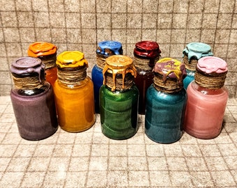 May include: A collection of nine small glass bottles filled with shimmering colored liquids. Each bottle features a cork stopper sealed with decorative wax in a matching or contrasting color, and the neck is wrapped in brown twine.
