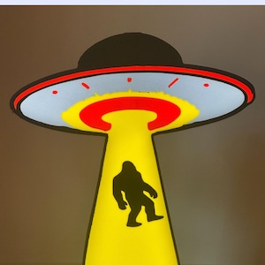 May include: A 3D-printed lightbox in the shape of a UFO, featuring a black silhouette of Bigfoot. The UFO has a black top with a red and white trim, a light blue underside, and a yellow beam. The text "Digital Download 3d Print Lightbox" is at the top.