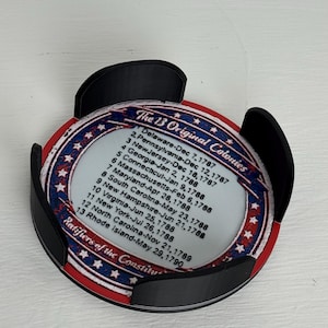 May include: A round coaster with a patriotic design featuring the text "The 13 Original Colonies" and a list of dates. The coaster is held in a black holder. The design includes red, white, and blue colors.