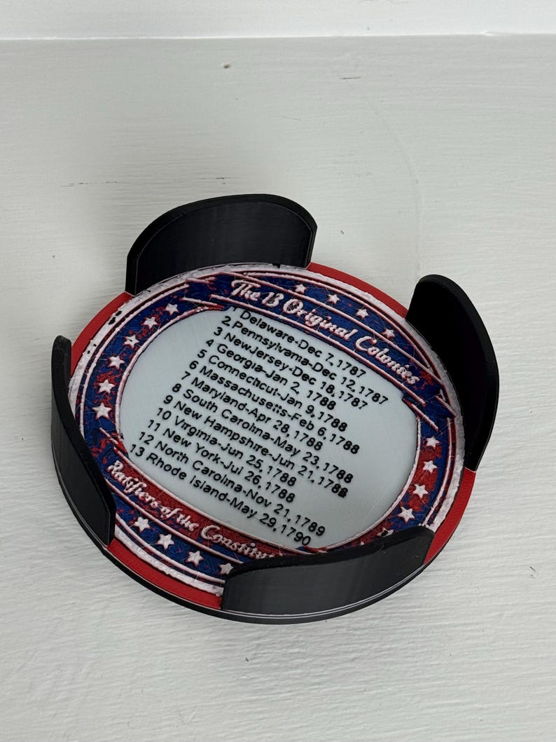May include: A round coaster with a patriotic design featuring the text "The 13 Original Colonies" and a list of dates. The coaster is held in a black holder. The design includes red, white, and blue colors.