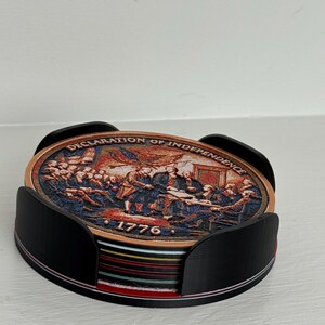 May include: A set of coasters featuring a depiction of the signing of the Declaration of Independence, with the text "DECLARATION OF INDEPENDENCE" and "1776". The coasters are held in a black holder.
