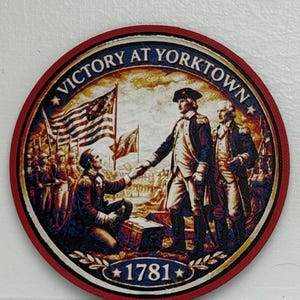 May include: Round wooden coaster with a red border featuring a historical illustration of the Victory at Yorktown. The image includes the text "Victory at Yorktown" and "1781", along with the word "YorkTown" at the top.