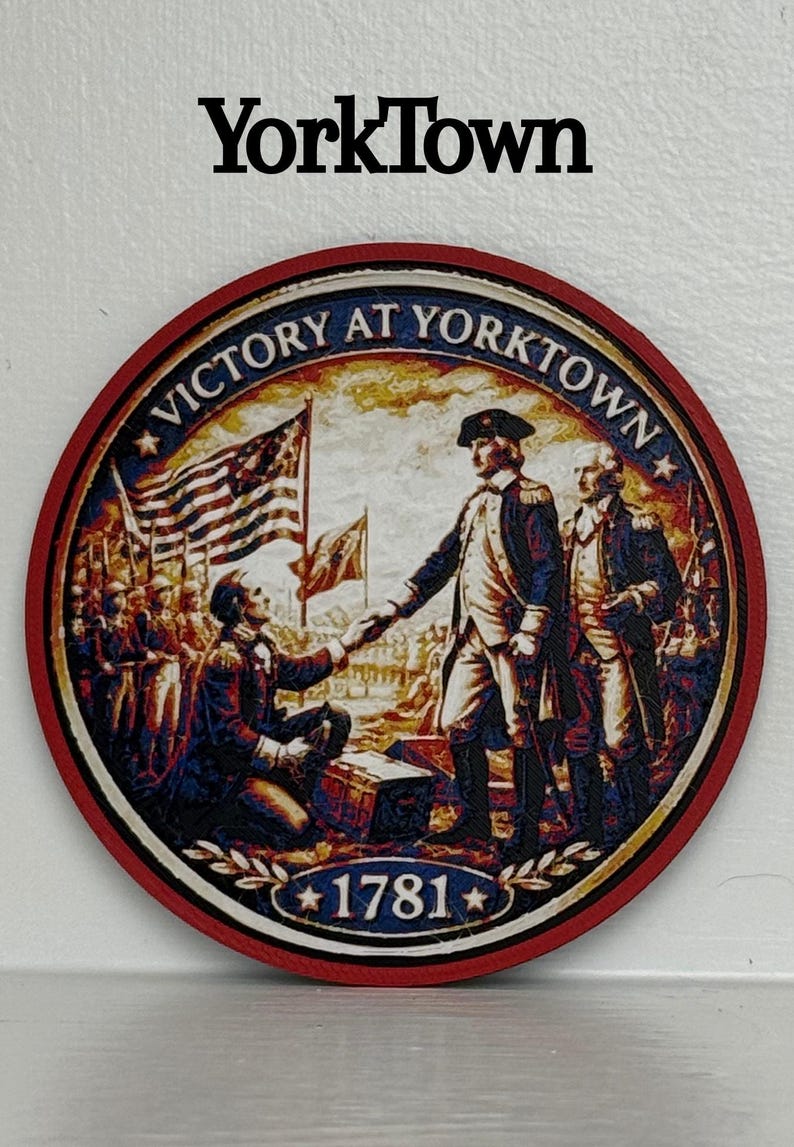 May include: Round wooden coaster with a red border featuring a historical illustration of the Victory at Yorktown. The image includes the text "Victory at Yorktown" and "1781", along with the word "YorkTown" at the top.
