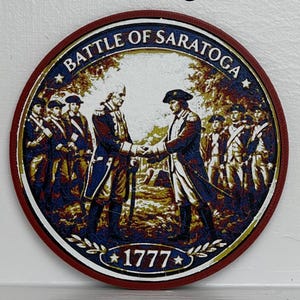 May include: Circular emblem with the text "Saratoga" at the top. The emblem features an illustration of two men shaking hands, surrounded by other figures. The words "BATTLE OF SARATOGA" and the year "1777" are also present.