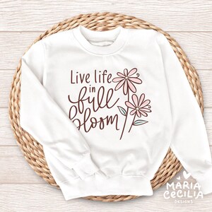 May include: White sweatshirt with the text "Live life in full bloom" in a brown script font. The design includes two pink flowers with green and blue accents. The sweatshirt is displayed on a woven, circular surface.