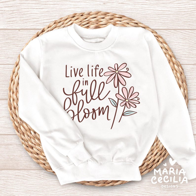 May include: White sweatshirt with the text "Live life in full bloom" in a brown script font. The design includes two pink flowers with green and blue accents. The sweatshirt is displayed on a woven, circular surface.