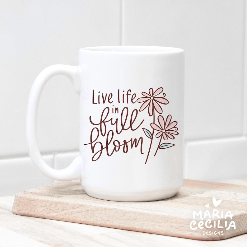 May include: White ceramic mug with a curved handle. The mug features the phrase "Live life in full bloom" in brown script, accompanied by pink flower illustrations. The mug is placed on a wooden cutting board.