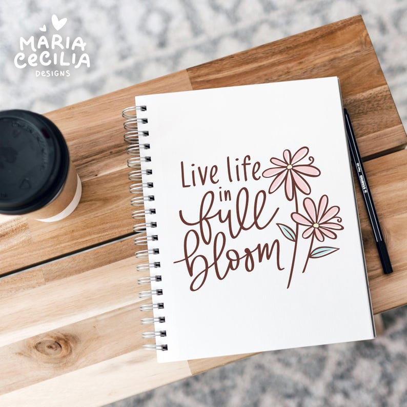 May include: A white spiral-bound notebook with the words "Live life in full bloom" in brown script, accompanied by pink flower illustrations. A black pen and a coffee cup sit nearby on a wooden surface. The notebook is from Maria Cecilia Designs.