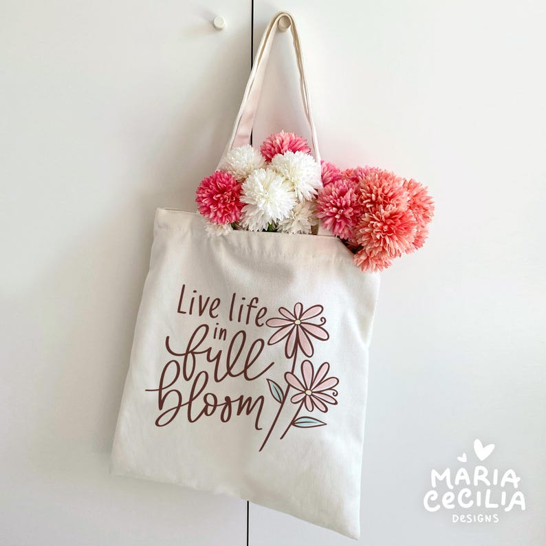 May include: A cream-colored tote bag with the phrase "Live life in full bloom" in brown script and floral illustrations. The bag is filled with pink and white artificial flowers, hanging against a white background.