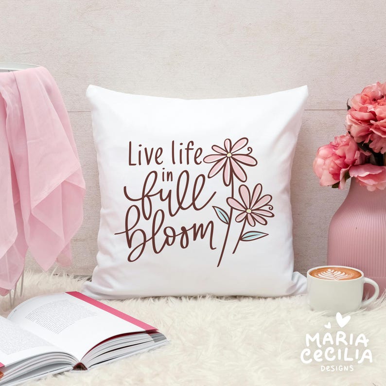 May include: A white square pillow with the phrase "Live life in full bloom" in brown script and floral illustrations. The pillow is on a white fluffy surface, next to a pink vase with flowers and a cup of coffee.