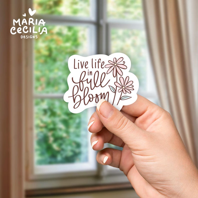 May include: A white sticker with the text "Live life in full bloom" in a handwritten font, accompanied by two pink flower illustrations. The sticker is held in a hand, with the Maria Cecilia Designs logo in the upper left corner.
