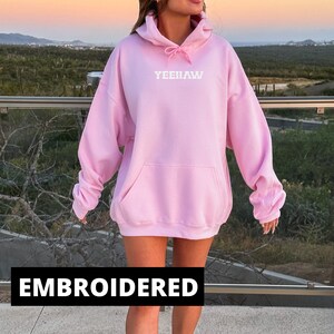 May include: A pink oversized hoodie with a white embroidered "YEEHAW" on the front. The hoodie has a drawstring hood and a kangaroo pocket.