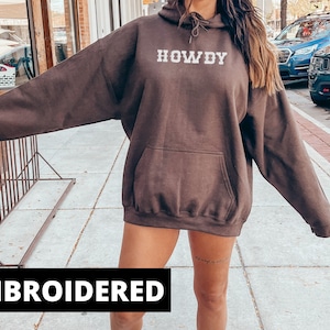 May include: A brown hoodie with a large white embroidered word "HOWDY" on the front. The hoodie has a kangaroo pocket and a drawstring hood.