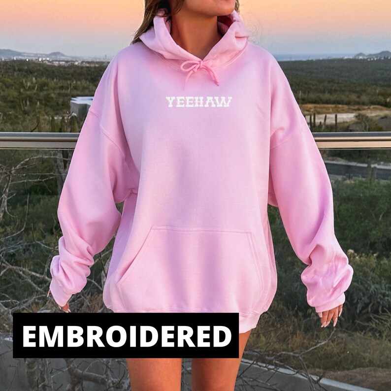 May include: A pink oversized hoodie with a white embroidered "YEEHAW" on the front. The hoodie has a pouch pocket and a drawstring hood.