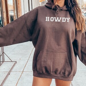 May include: A brown hoodie with a white "HOWDY" graphic on the front. The hoodie has a drawstring hood and a kangaroo pocket.