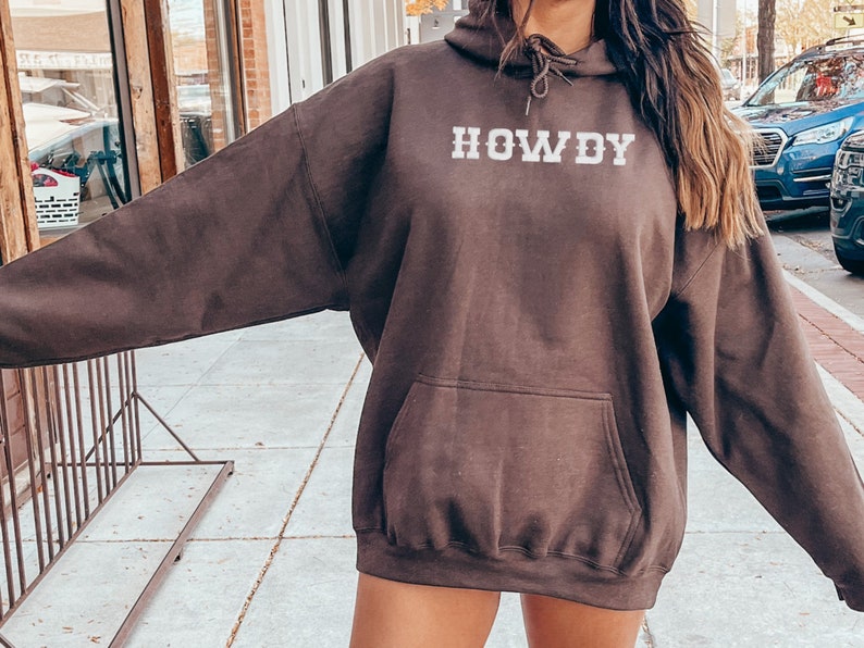 May include: A brown hoodie with a white "HOWDY" graphic on the front. The hoodie has a drawstring hood and a kangaroo pocket.
