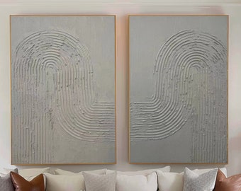 Puede incluir: Dos pinturas abstractas con un fondo blanco texturizado y líneas grises que forman un patrón curvo. Las pinturas están colgadas en una pared blanca sobre un sofá seccional blanco con almohadas marrones. Una mesa de café de madera con libros y un jarrón de flores blancas se encuentra frente al sofá.