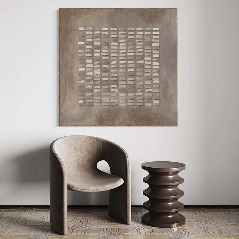 May include: A brown abstract painting with a grid pattern of white and gray squares hangs on a white wall above a brown chair and a brown side table.
