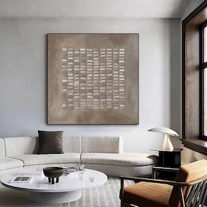May include: A brown and white abstract painting with a grid pattern of rectangles. The painting is hanging on a white wall in a living room setting with a white couch, a coffee table, and a chair.