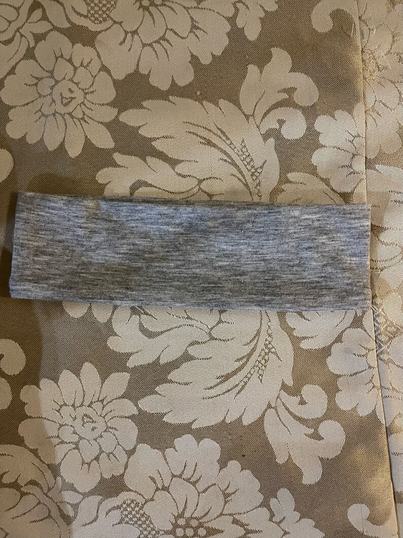 May include: A gray headband made of a soft, stretchy fabric. The headband is folded in half and lying flat on a patterned surface.