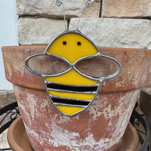 May include: A stained glass bee with yellow and black stripes, with a metal frame, is attached to a terracotta flower pot.