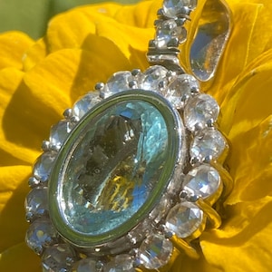 May include: An oval-shaped pendant featuring a large, faceted light blue central gemstone surrounded by a halo of smaller, clear, round-cut stones. The pendant includes a metallic bail at the top, also adorned with a vertical row of clear stones.