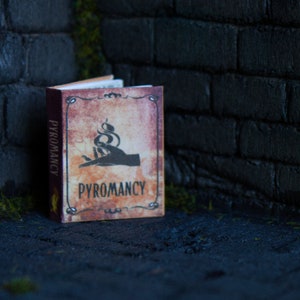 Puede incluir: Un libro en miniatura titulado "Pyromancy" con una portada de estilo vintage. La portada presenta una mano sosteniendo una serpiente con llamas, y el título está escrito en una fuente decorativa. El libro está colocado contra una pared de ladrillos oscuros.