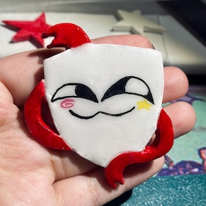 Puede incluir: Un pin hecho a mano en forma de escudo blanco con una cara de dibujos animados, ojos negros y un rubor rosa. Tiene brazos rojos y una cresta roja. Una estrella amarilla está pintada en el lado derecho de la cara. El pin se sostiene en una mano.