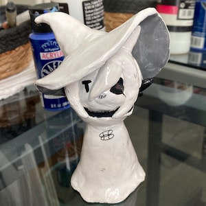 Puede incluir: Una figura de cerámica blanca de un personaje que lleva un sombrero de bruja con ala gris. El personaje tiene una "T" negra en la cara, ojos negros y una boca sonriente. La figura tiene un cuerpo en forma de campana y un símbolo negro en el frente.